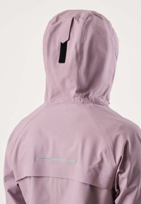Osoba v lehce fialové kapuci viděná zezadu, s viditelnými šitými švy a reflexní značkou značky na horní části zad.