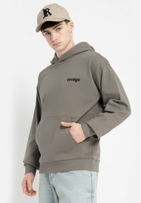 Młody mężczyzna w szarej bluzie z kapturem z napisem "ravage", jasnoniebieskich dżinsach i beżowej czapce z daszkiem z czarnym logo "R".