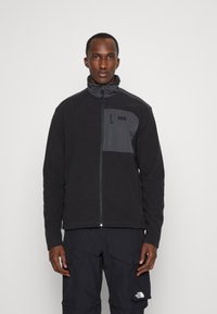 Veste en polaire noire avec fermeture éclair sur le devant, col montant et poche poitrine. Panneau d'accent gris en haut à gauche. Texture douce et design décontracté.