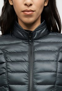 Μαύρο μπουφάν puffer με ψηλό γιακά, έντονη ραμμένη υφή και εμπρόσθιο φερμουάρ. Ομαλή, γυαλιστερή επιφάνεια, μίνιμαλ σχεδιασμός.