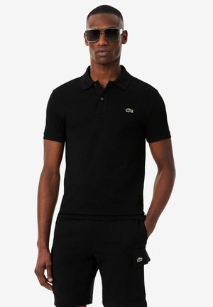 Uomo con polo nera e pantaloncini cargo con toppe logo, in piedi con una mano in tasca, indossa grandi occhiali da sole scuri.