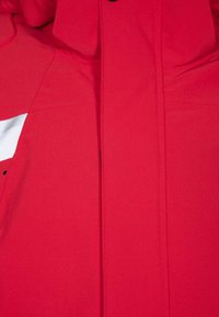 Chaqueta roja con una textura suave y mate, con un acento blanco en el lado izquierdo y botones negros a lo largo del cierre frontal.