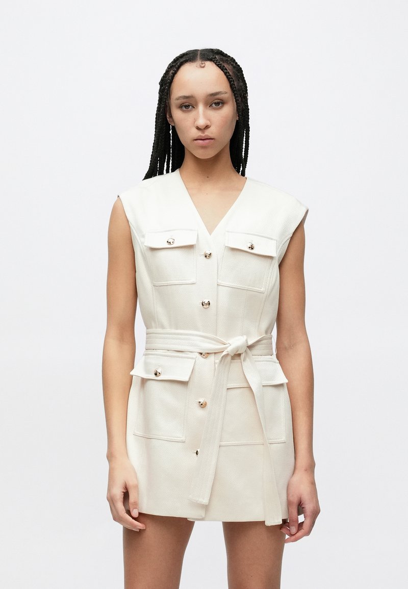 Μπεζ αμάνικο φόρεμα με v-λαιμόκοψη, χρυσά κουμπιά διακοσμητικά, τέσσερις τσέπες και δεμένη ζώνη. Υφή υφάσματος, μήκος μέχρι το γόνατο.