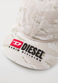 Căciulă de camuflaj cu modele bej și verde deschis. Prezintă un logo "DIESEL DENIM DIVISION" în roșu și negru pe partea din față. Fabricată din material textil.