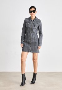 Grå denimkjole med lange ermer, glidelås foran, krage og to brystlommer. Brukt med svarte ankelstøvler og store svarte solbriller.