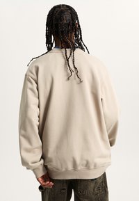 Beige oversized sweatshirt made of soft fabric, featuring a round neckline, ribbed cuffs, and a simple, smooth texture. Back view shown.