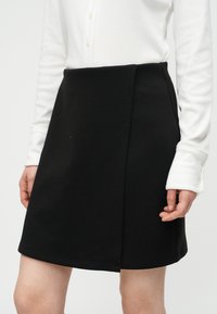 Γυναίκα που φοράει μαύρη φούστα γραμμής Α με λεπτομέρεια τύλιγμα και λευκό μακρυμάνικο πουκάμισο με κουμπιά, ορατή από τη μέση έως το μέσο του μηρού.