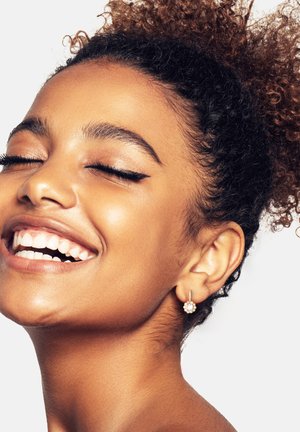 Primo piano di un volto sorridente con capelli ricci, che indossa un orecchino d'argento con un design floreale e un gruppo di piccole pietre rotonde.