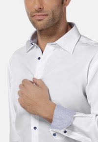 Witte shirt met een gestructureerde kraag. De manchetten hebben een blauw geometrisch patroon. Blauwe knopen contrasteren met de strakke stof van het shirt.
