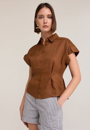Frau mit kurzen braunen Haaren, die ein braunes kurzärmliges Hemd mit Knöpfen und gestreifte Shorts trägt, mit einer Hand in der Tasche.