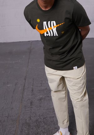 Homme portant un t-shirt noir Nike Air avec un swoosh orange, un pantalon beige et des baskets blanches, se tenant sur un sol en béton gris.