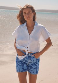 Valkoinen pellavainen lyhythihainen paita, solmittu vyötärölle, yhdistetty sinisiin kuvioituihin farkkushortseihin, joissa on kukkakuosi ja hiekkainen rantatausta.