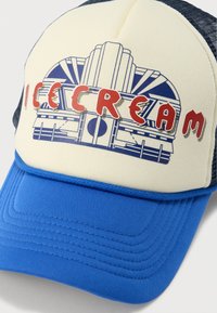 Καπέλο μπέιζμπολ με κρεμ πίνακα στο μπροστινό μέρος που απεικονίζει ένα μπλε γραφικό καταστήματος παγωτού και κόκκινα γράμματα. Μπλε ύφασμα στο γείσο και πλέγμα στην πίσω πλευρά.