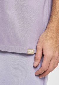 Jasnofioletowa koszulka wykonana z miękkiego materiału, z przyszytą metką z napisem "DEF." na dolnym brzegu, zbliżenie na rękę trzymającą materiał.