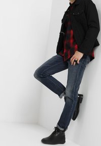 Svart denimjacka över en röd och svart rutig skjorta, i kombination med blå jeans och svarta ankelstövlar, stående mot en vit bakgrund.