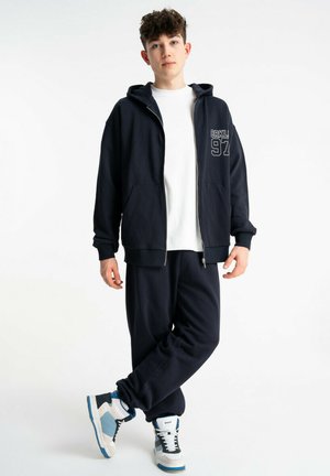 Nastolatek stojący w granatowej bluzie na zamek z napisami "BRKLYN 97", dresowych spodniach do kompletu, białej koszulce i biało-niebieskich trampkach na białym tle.