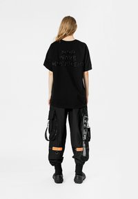 Camiseta negra con "MOVIMIENTO MOD WAVE" en letras texturizadas en la parte posterior, combinada con pantalones cargo negros holgados que cuentan con múltiples bolsillos y acentos.
