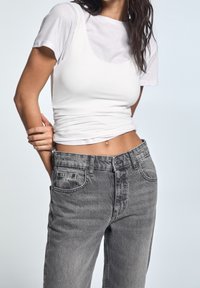 Bílý přiléhavý top bez rukávů přetažený přes bílý tričko s krátkým rukávem, ve spojení se šedými džíny s vysokým pasem, které mají distressed vzhled a kapsy.