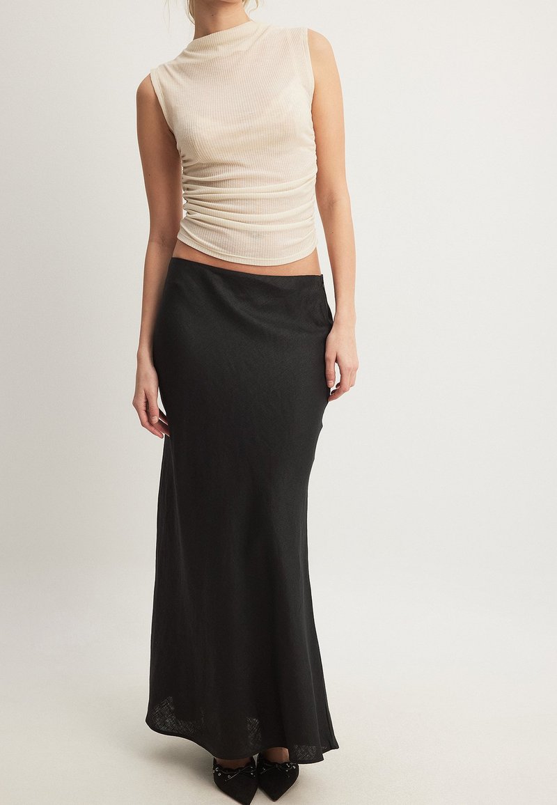 Top brez rokavov v bež barvi z naborki, vparen z dolgim črnim krilom z visokim pasom in črnimi koničastimi čevlji na svetli podlagi.