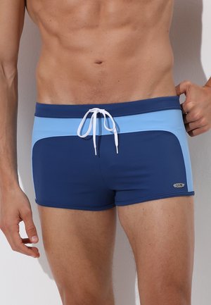 Torsetto maschile che indossa pantaloncini da bagno blu navy e azzurro chiaro con coulisse bianca, in piedi con una mano che sistema la vita.