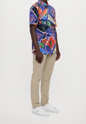 Kurzärmeliges Hemd mit Knöpfen aus mehrfarbiger Baumwolle mit verschiedenen Sportgrafiken, kombiniert mit beige Hosen und weißen Turnschuhen.