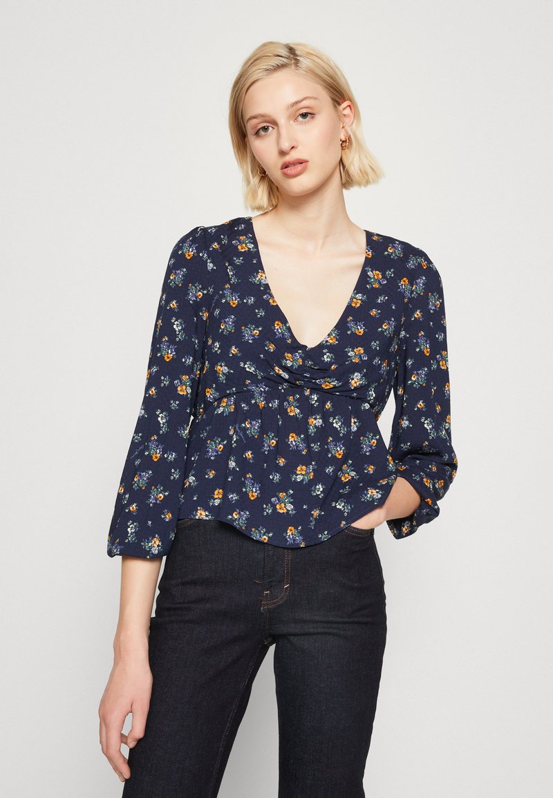Blusa floreale navy con scollatura a V profonda, maniche a sbuffo e vita arricciata. Presenta piccoli fiori colorati su un tessuto testurizzato.