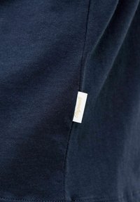 Σκούρο ναυτικό μπλε ύφασμα με μια μικρή λευκή ετικέτα που γράφει «Originals» ραμμένη στην πλευρική ραφή του ρούχου.
