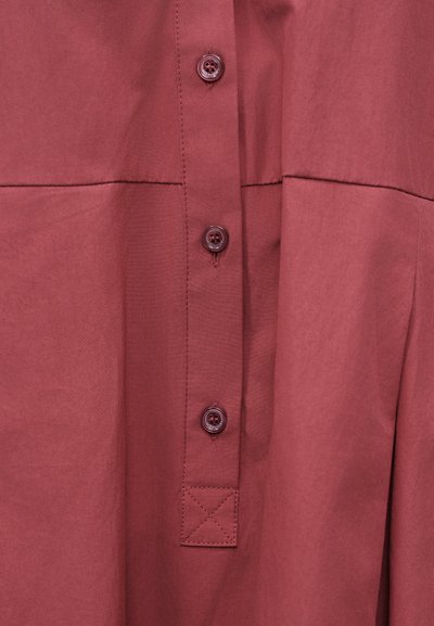 Κοντινή λήψη σκούρου κόκκινου υφάσματος με τρία κουμπιά στο ίδιο χρώμα, τοποθετημένα κάθετα σε ένα ραμμένο λάστιχο με λεπτομερή ραφή και τετράγωνο ραμμένο μπαλώμα.