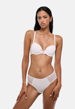 Femme portant un soutien-gorge blanc et une culotte en mesh transparent assortie sur un fond uni clair, debout, les mains le long du corps.