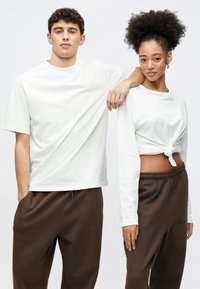 Άνδρας με λευκό βαμβακερό t-shirt με στρογγυλή λαιμόκοψη. Γυναίκα με λευκή μακρυμάνικη μπλούζα δεμένη στη μέση, συνδυασμένη με καφέ παντελόνι.