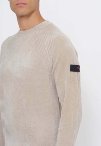 Beiger, strukturierter Strickpullover mit einem Rippenmuster an den Schultern und einem schwarzen Stoffpatch mit Knöpfen am linken Ärmel.