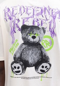 Weiße T-Shirt mit einem Grafik eines grauen Bären mit grünen Akzenten und gestickten Gesichtszügen, überlagert mit lila Text und neongrünen Details.