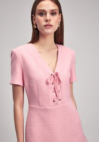 Rosa strukturiertes Kleid mit kurzen Ärmeln, V-Ausschnitt und geschnürtem Frontbereich mit metallenen Ösen und Ripsbandbindungen. Minimalistisches, figurbetontes Design.