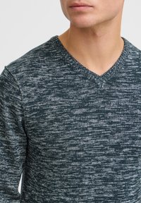 Mørkegrønn og grå strikket genser med V-hals-design. Stoffet har et teksturert mønster med et subtilt, blandet utseende.