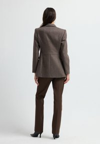 Blazer marrone su misura con texture a spina di pesce, chiusura frontale con un bottone, rever affilati e silhouette aderente, abbinato a pantaloni dritti marroni.