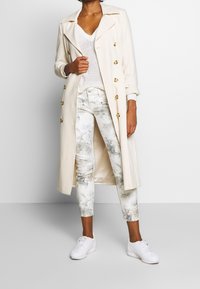 Długi kremowy trencz z złotymi guzikami, zestawiony z białym swetrem i dżinsami o skinny kroju w kwiatowy wzór. Całość dopełniają białe sneakersy.