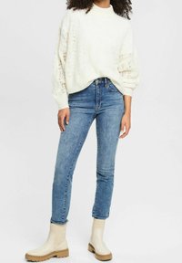Crème kleurige, kabelgebreide trui met een hoge hals en ballonnenmouwen, gecombineerd met blauwe skinny jeans en beige enkellaarsjes met een dikke zool.