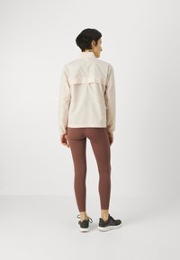 Giacca leggera rosa chiaro con maniche lunghe, dettaglio sulla parte posteriore, abbinata a leggings marroni e scarpe da ginnastica nere.
