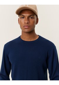 Marineblauer Strickpullover mit langen Ärmeln und geripptem Kragen. Mit subtiler Logo auf der Brust. Das Model trägt eine beige Mütze.