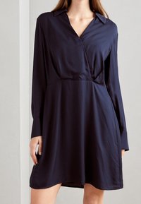 Marineblaues Kleid aus glattem Stoff mit Kragen, langen Ärmeln und taillierter Taille, das in einen knielangen Rock übergeht.