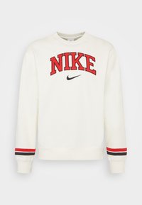 Krämfärgad sweatshirt med röd broderad text "NIKE". Har en liten svart Nike-logotyp. Ribbad muddar med röda och svarta ränder. Klassisk passform.