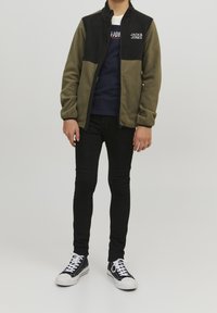 Veste en polaire avec un design en blocs de couleurs noir et vert olive, fermeture éclair, et logo sur la poitrine, associée à un jean slim noir et des baskets noires.