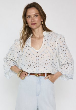 Femme aux cheveux châtain clair porte un chemisier blanc en dentelle œillet à manches 3/4, un jean taille haute bleu clair, des boucles d'oreilles créoles dorées et des bracelets.
