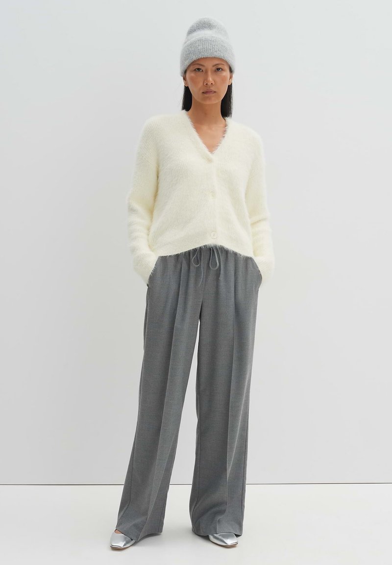 Cardigan fluffy crema con scollo a V e chiusura con bottoni, abbinato a pantaloni grigi con coulisse e scarpe slip-on argento. Cappello beanie grigio.