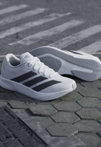 Chaussures de course Adidas blanches avec des bandes noires posées sur un pavement extérieur texturé près d'un passage piéton.