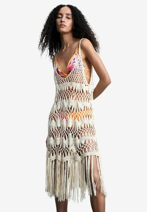 Femeie cu păr creț purtând un bikini colorat sub o rochie tip plasă croșetată crem, lungă până la genunchi, cu franjuri la tiv.