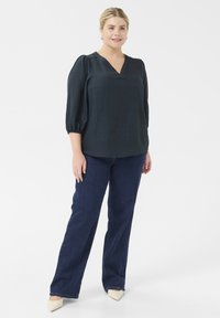 Donkergroene, gestructureerde blouse met een V-hals en driekwart pofmouwen, gecombineerd met donkerblauwe, rechte jeans en beige blokhak schoenen.