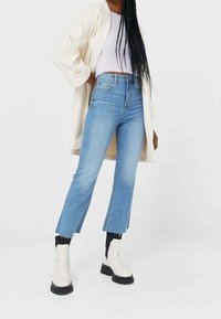 Light blue high-waisted jeans with a straight leg and frayed hem, paired with a white cropped top and cream coat; chunky white ankle boots.