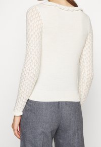 Kremowy sweter z falbankową linią szyi; rękawy z koronkowym wzorem; dzianinowa faktura; dopasowana talia; zestawiony z szarymi spodniami.