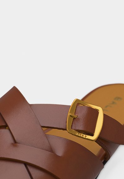 Lanière de sandale en cuir marron avec boucle carrée dorée, montrant les détails de texture et de couture sur un fond beige.